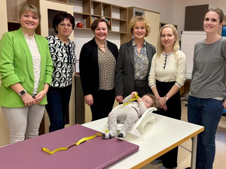 Fünf Frauen in Berufskleidung stehen um einen Tisch mit einem Baby darauf. Das Baby sitzt in einem Babysitz und hält ein Maßband. Die Frauen lächeln.
