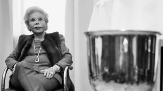 Eine ältere Frau mit Brille und einer Halskette sitzt auf einem Stuhl mit den Händen auf dem Schoß. Sie scheint für ein Foto zu posieren.