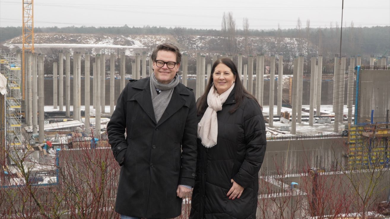 Ein Mann und eine Frau stehen nebeneinander in Winterkleidung. Sie lächeln und posieren für ein Foto. Hinter ihnen befindet sich eine Baustelle mit einer Wand und Metallpfählen.