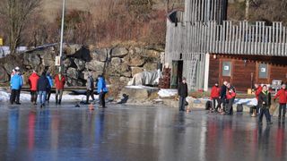 Mehrere Menschen stehen auf einer Eisbahn mit orangefarbenen Kegeln, einige in Jacken und Stiefeln. Dahinter steht ein Holzgebäude mit einer Leiter.