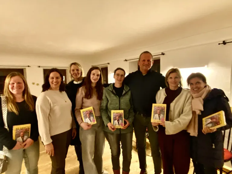 Sieben Erwachsene stehen in einem Raum und halten Bücher in den Händen. Ein Mann in der Mitte hat seine Arme um zwei Frauen gelegt. Die Bücher tragen den Titel 'SO GEHT'S'.