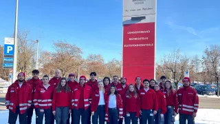 Eine Gruppe von Menschen in roten Uniformen und roten Mützen steht vor einem großen Schild des Roten Kreuzes mit verschneirtem Hintergrund.