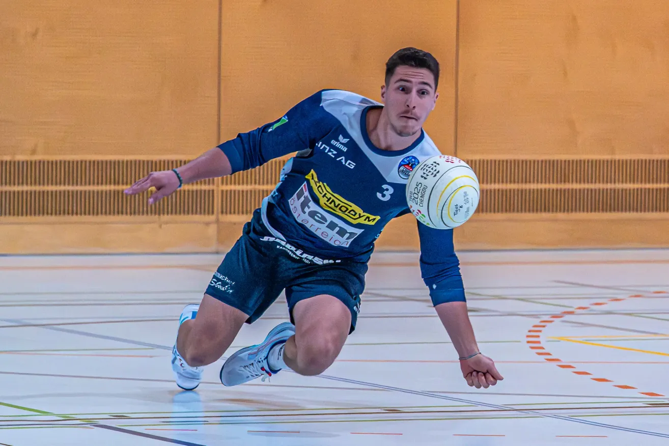 Ein Mann in blauer Sportbekleidung rutscht auf dem Spielfeld und fängt einen Ball. Er trägt das Trikot mit der Nummer 3 und ein Armband. Der Ball hat ein gelbes und weißes Design.