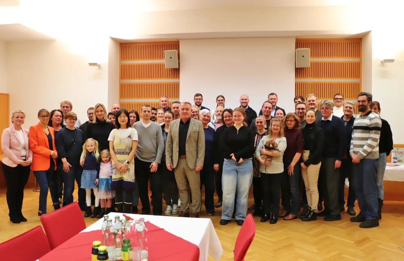 Eine Gruppe von Menschen posiert für ein Foto in einem Raum mit Holzwänden und einem Tisch mit Flaschen. Einige tragen Brillen, andere haben Haustiere.