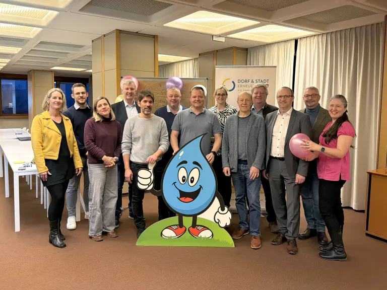 Eine Gruppe von Menschen steht in einem Innenraum, wahrscheinlich einem Konferenzraum, in formeller Kleidung. In der Mitte ist eine große Cartoon-Figur zu sehen. Ein Banner ist hinter ihnen angebracht.