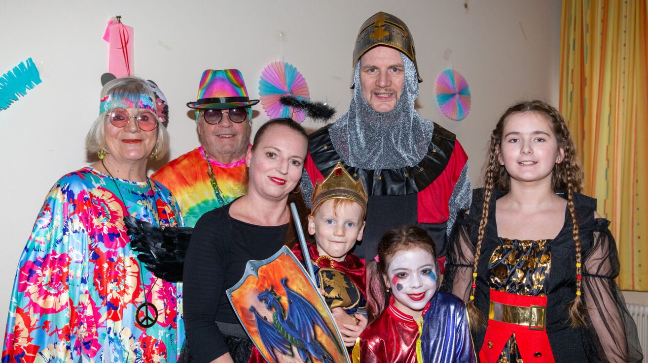 Eine Familie in farbenfrohen Kostümen, wahrscheinlich für eine thematische Veranstaltung. Eine Frau hält einen Drachenschild, und ein Junge und ein Mädchen tragen Ritter- und Clownkostüme.