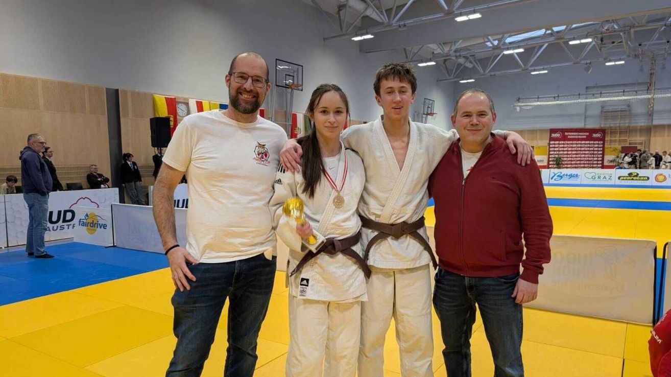 Vier Personen stehen gemeinsam auf einem gelben Teppich und feiern wahrscheinlich einen Kampfsporterfolg. Die beiden in der Mitte tragen weiße Uniformen, einer hält einen goldenen Pokal. Die anderen, in Freizeitkleidung, lächeln und posieren für ein Foto. Im Hintergrund ist ein Basketballkorb zu sehen.