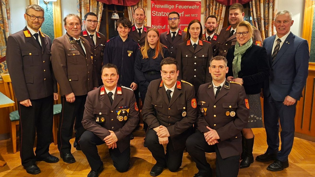 Eine Gruppe von Feuerwehrleuten in Uniform, einige kniend, andere stehend, posiert für ein Foto vor einem roten Banner.