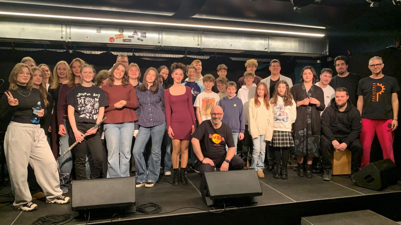 Eine Gruppe von Menschen, möglicherweise ein Theaterensemble, posiert auf einer Bühne mit zwei Lautsprechern und lächelt für ein Foto. Ein Mann in einem Hard Rock Cafe-Shirt kniet.