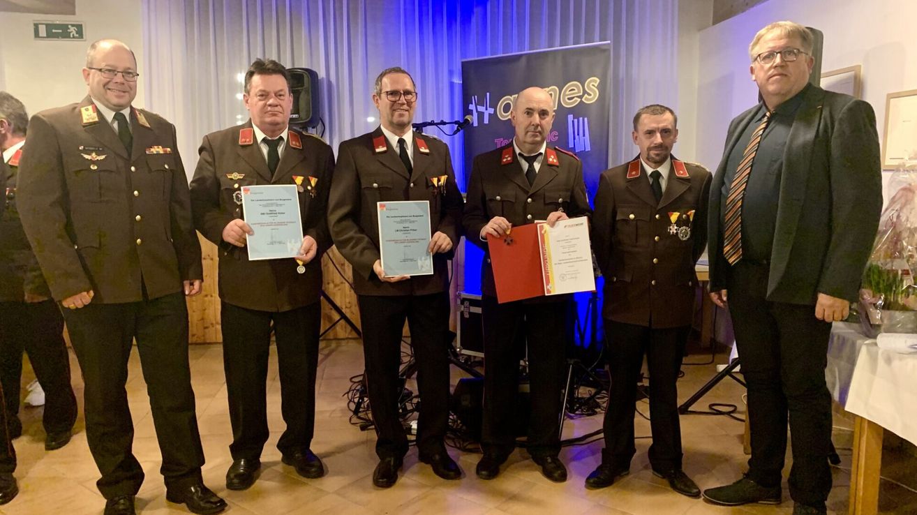 Vier Männer in Uniformen stehen zusammen und halten Urkunden und Medaillen. Sie stehen vor einem Banner mit einem Logo und Text. Dahinter befindet sich eine Bühnenausstattung mit Mikrofonen, Lautsprechern und einem Lautsprecherständer.