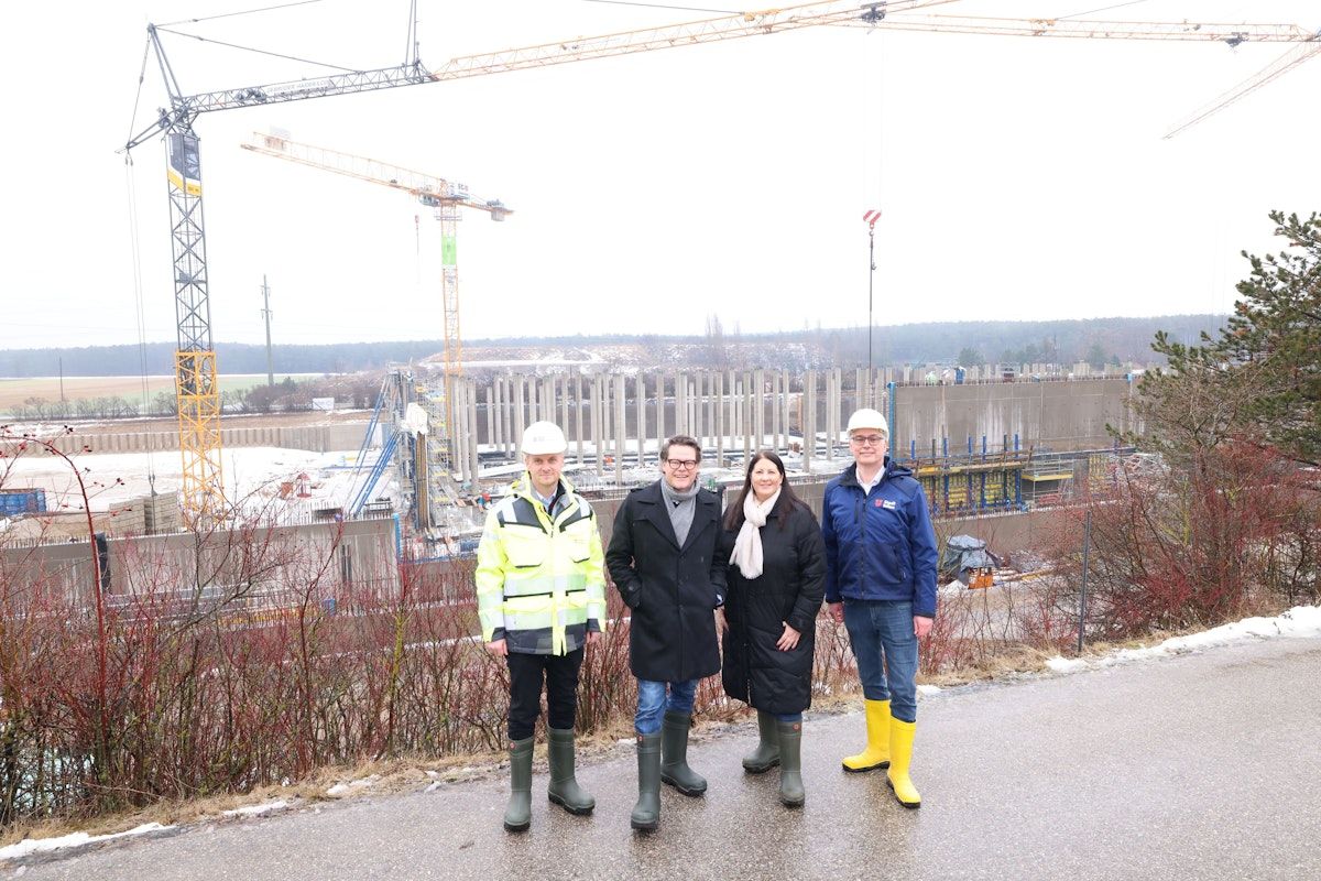 Vier Personen in Warnwesten und Stiefeln stehen vor einer Baustelle. Sie tragen Winterkleidung und Schutzhelme. Dahinter wird eine Betonmauer gebaut. Kräne sind im Hintergrund sichtbar.