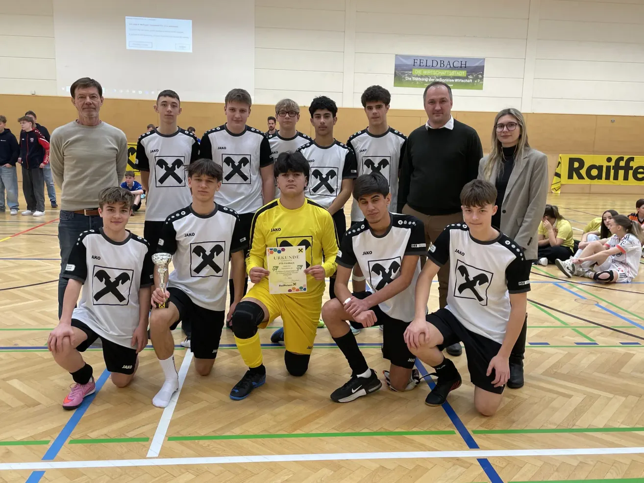 Eine Fußballmannschaft posiert mit einem Pokal für ein Foto. Die Mannschaft trägt einheitliche Trikots, und ein Mann hält eine Urkunde. Hinter ihnen ist eine Fahne an der Wand angebracht.