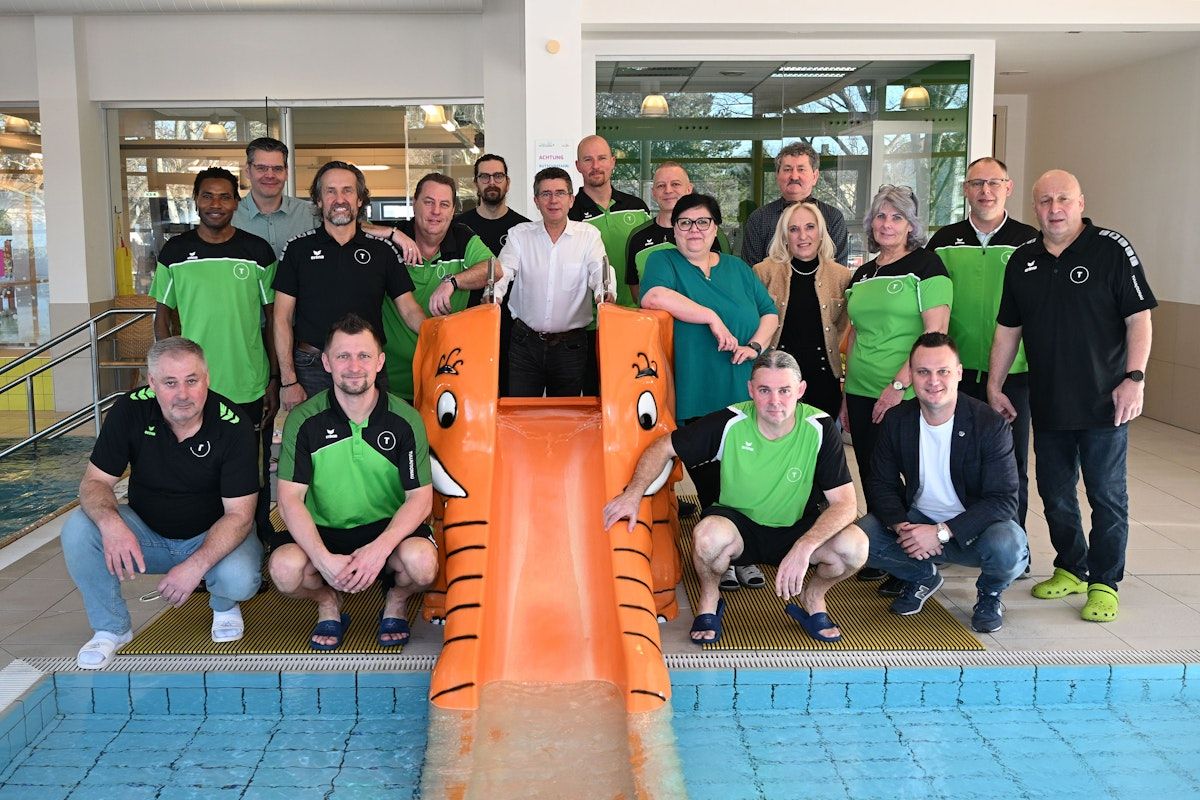 Eine Gruppe von Erwachsenen, einige mit Brille, posiert für ein Foto vor einem Pool mit einem Elefanten geformten Rutschen. Sie tragen alle grüne und schwarze Kleidung.