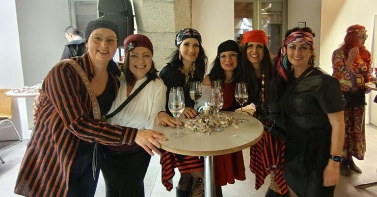 Eine Gruppe von Frauen in Piratenkostümen posiert für ein Foto um einen Tisch mit Weingläsern und Snacks und lächelt fröhlich.