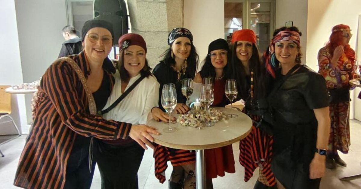 Eine Gruppe von Frauen in Piratenkostümen posiert für ein Foto um einen Tisch mit Weingläsern und Snacks und lächelt fröhlich.
