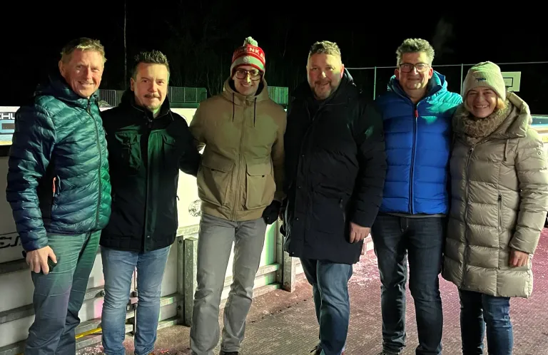 Vier Männer stehen zusammen auf einer Eisbahn bei Nacht und lächeln für ein Foto. Sie tragen verschiedene Jacken, einer trägt eine Mütze.