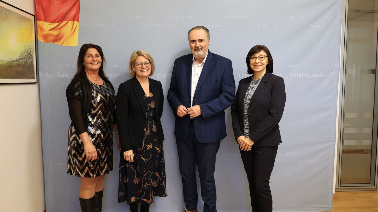 Fünf Personen stehen vor einer Wand mit Text und lächeln für ein Foto. Der Mann in der Mitte trägt einen blauen Anzug. Zwei Frauen an den Seiten tragen schwarze Blazer. Der Hintergrund hat verschwommenen Text.