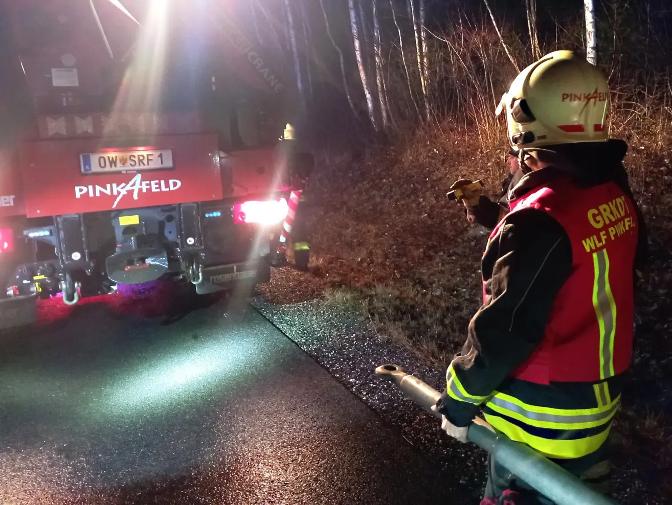 Ein Feuerwehrmann steht neben einem Fahrzeug mit der Aufschrift 'PINKFELD' in einem dunklen Bereich. Die Rückseite des Fahrzeugs ist beleuchtet, und der Feuerwehrmann hält ein Werkzeug. Die Szene ist von Bäumen umgeben.