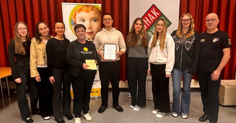 Eine Gruppe von Menschen steht lächelnd zusammen. Ein Mann hält ein Zertifikat und ein gelbes Papier. Sie stehen vor einer Kulisse mit Logos und einem Poster eines Kindes.