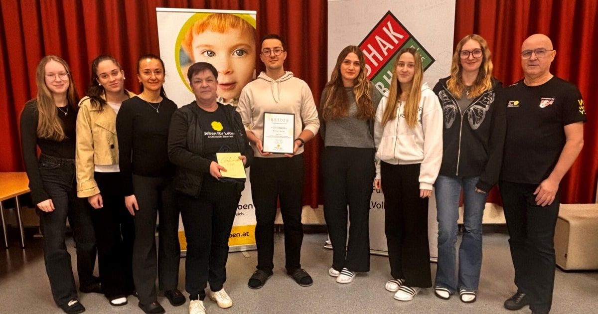 Eine Gruppe von Menschen steht lächelnd zusammen. Ein Mann hält ein Zertifikat und ein gelbes Papier. Sie stehen vor einer Kulisse mit Logos und einem Poster eines Kindes.