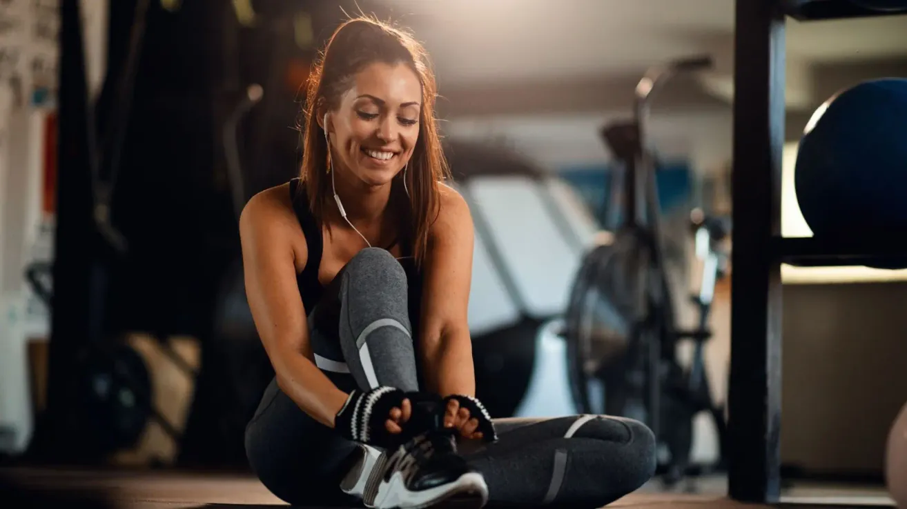 Eine lächelnde Frau mit Kopfhörern, in Trainingskleidung, sitzt auf dem Boden in einem Fitnessstudio und streckt sich möglicherweise vor dem Training. Hinter ihr ist Fitnessgeräte zu sehen, darunter ein stationäres Fahrrad.