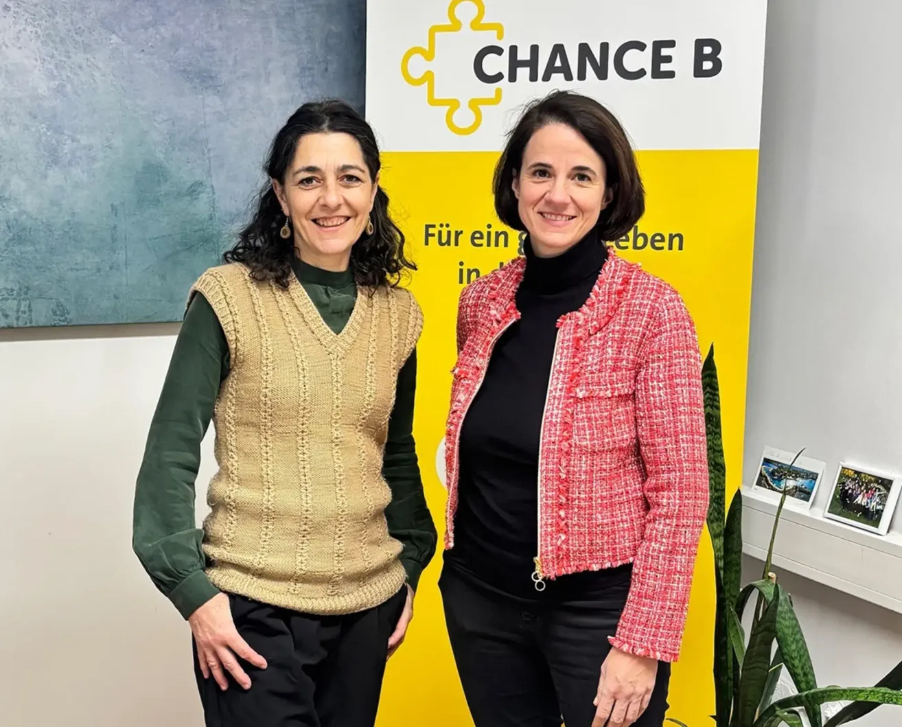 Zwei Frauen stehen vor einem 'Chance B'-Schild. Eine trägt eine beige Strickjacke, die andere eine rote Jacke. Beide lächeln.