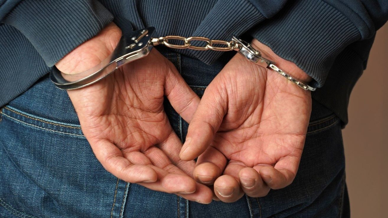 Nahaufnahme der Hände eines Erwachsenen mit silbernen Handschellen. Die Person trägt ein blaues Langarmshirt und Jeans.