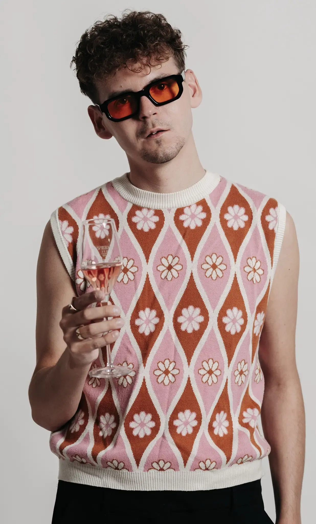 Ein Mann in einem ärmellosen Pullover mit geometrischem Blumenmuster hält ein Weinglas mit rosa Flüssigkeit. Er trägt eine Sonnenbrille und Ringe an den Fingern.