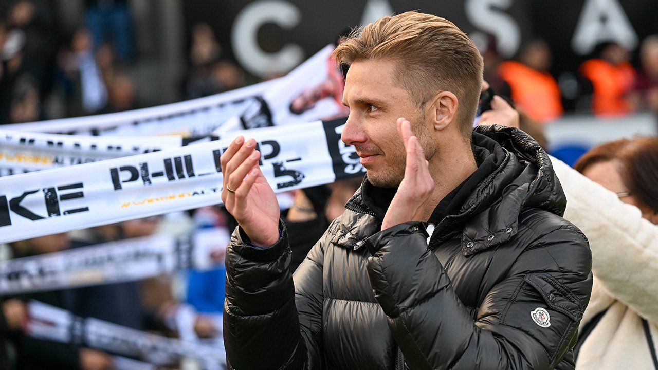 Ein Mann in einer schwarzen Daunenjacke klatscht mit den Händen vor einer Menge. Im Hintergrund sind verschwommene Menschen zu sehen, die Banner halten.
