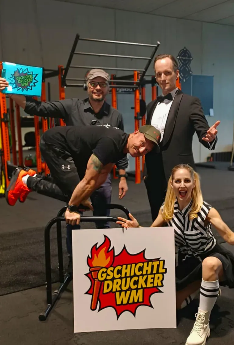 Vier Personen stehen in einem Fitnessstudio. Ein Mann lehnt sich mit den Füßen auf einem Ständer an eine Stange. Eine Frau kniet mit der Hand auf einem Schild, das 'Gschichtl Druckner' steht. Hinter ihnen zeigt ein Mann in einem Frack auf etwas. Ein weiterer Mann in einer Kappe steht neben ihnen.