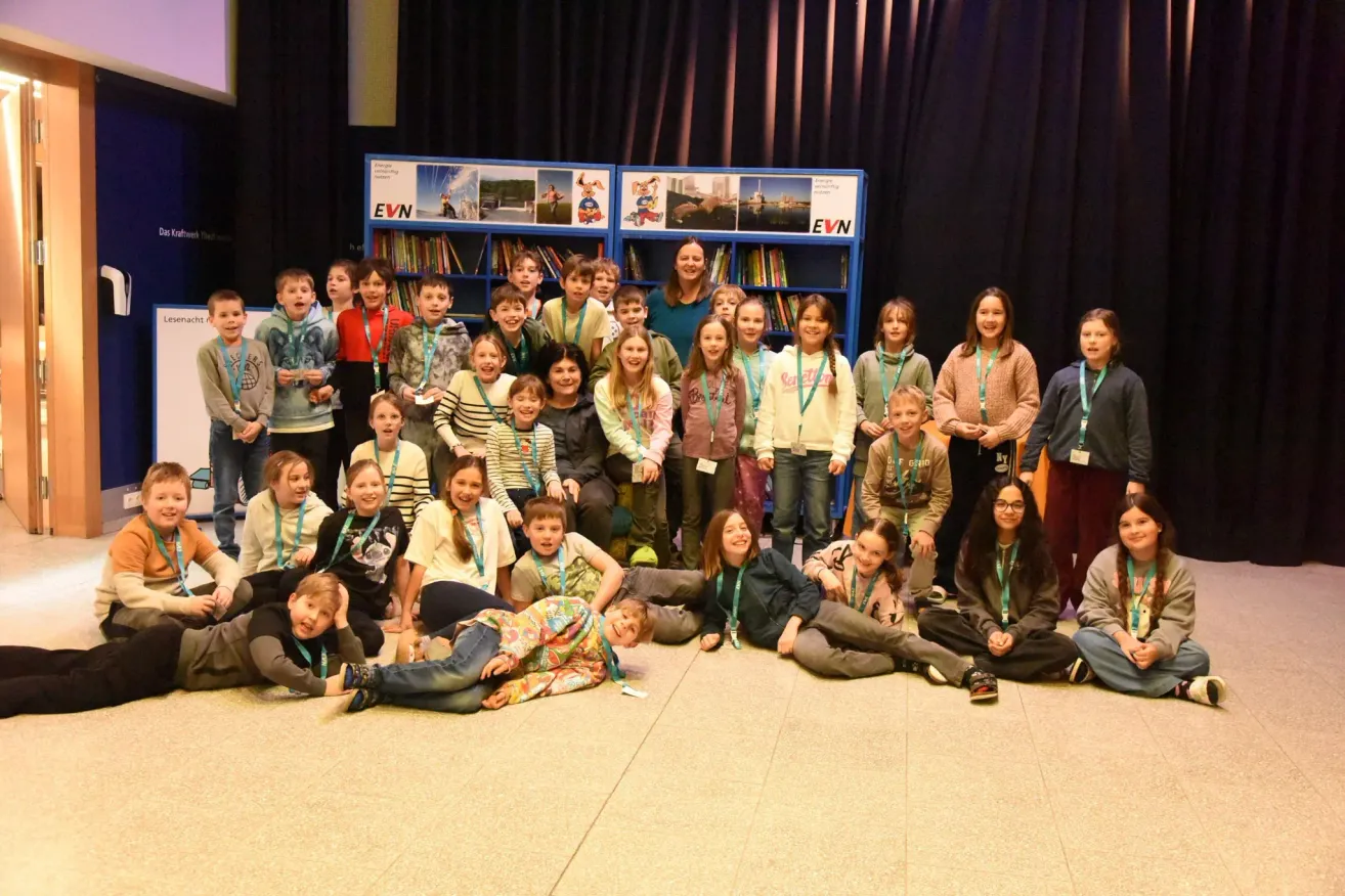 Eine Gruppe von Kindern und Erwachsenen versammelt sich in einem Raum mit blauen Regalen im Hintergrund, einige sitzen auf dem Boden, während andere stehen. Sie tragen alle grüne Lanyards und scheinen für ein Foto zu posieren.