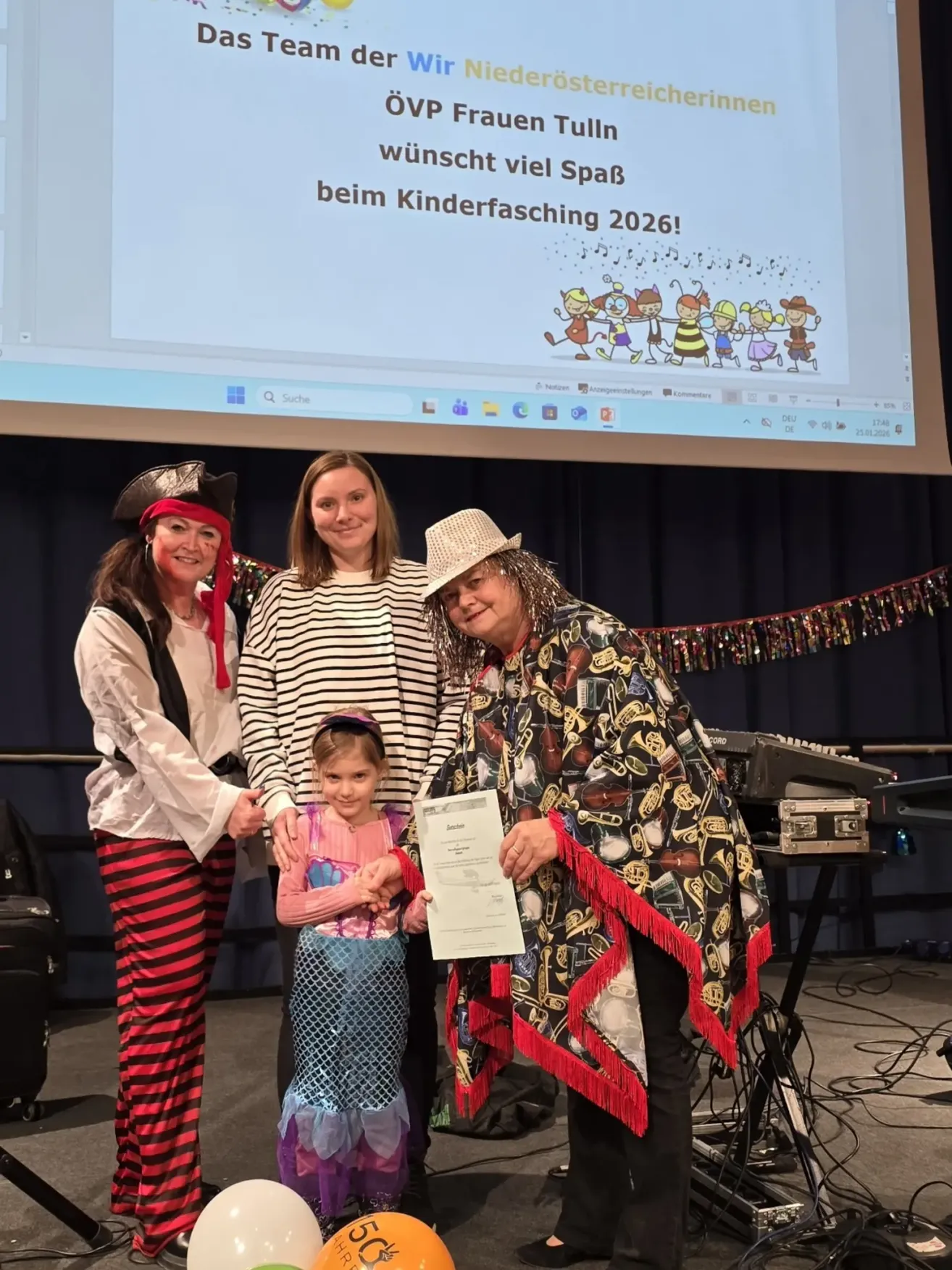 Drei Personen stehen auf einer Bühne mit einem Kind. Die Frau in der Mitte hält ein Zertifikat und lächelt. Die Frau auf der linken Seite ist als Piratin gekleidet, und die Frau auf der rechten Seite trägt ein farbenfrohes Outfit. Die Bühne hat eine Kulisse mit einer Projektionsleinwand, die eine Präsentation zeigt.