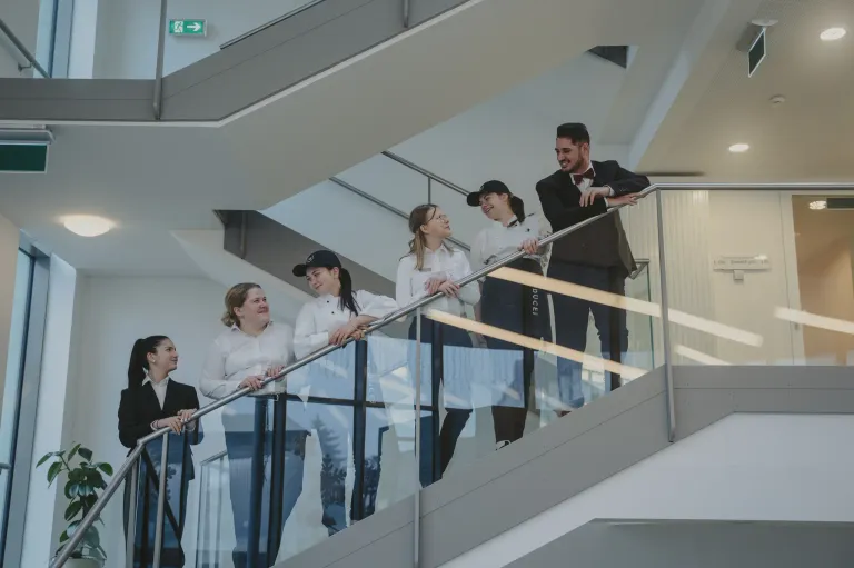 Eine Gruppe von Personen in weißen Kochuniformen steht auf einer Treppe und posiert für ein Foto. Sie lächeln und scheinen sich zu amüsieren. Die Treppe hat ein Glasgeländer und führt in eine höhere Etage.