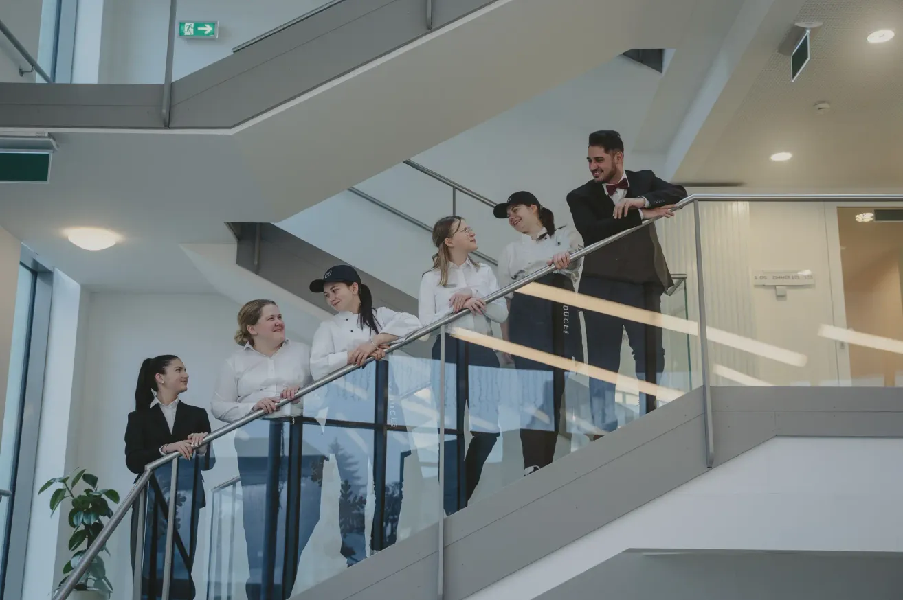 Eine Gruppe von Personen in weißen Kochuniformen steht auf einer Treppe und posiert für ein Foto. Sie lächeln und scheinen sich zu amüsieren. Die Treppe hat ein Glasgeländer und führt in eine höhere Etage.