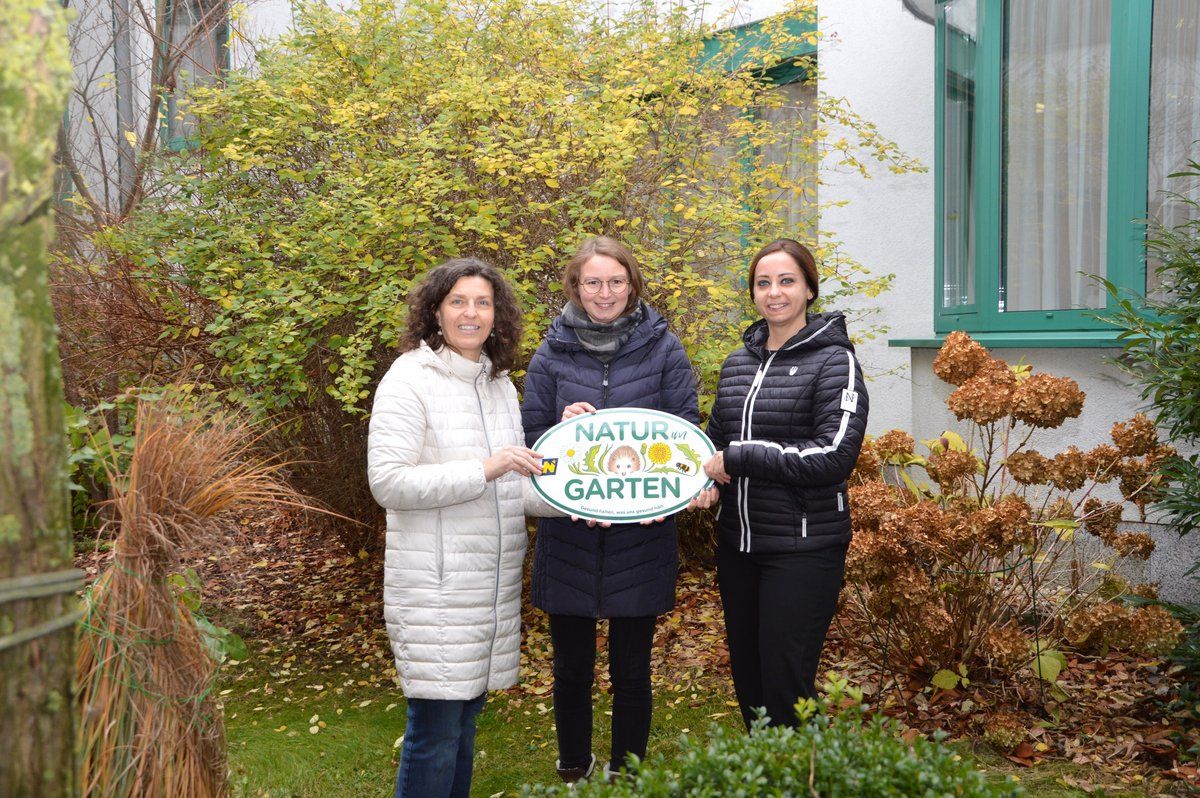Drei Frauen stehen vor einem Haus und halten ein grünes Schild mit der Aufschrift 'Natur im Garten'. Sie lächeln und scheinen für ein Foto zu posieren. Hinter ihnen befinden sich Büsche und ein Gebäude mit grünen Fensterrahmen.