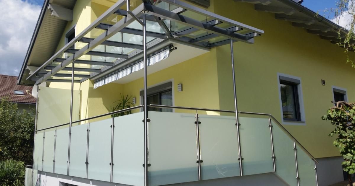 Ein Balkon mit Glasscheiben, einem Metallrahmen und einem versenkbaren Vordach ist an einem gelben Gebäude befestigt.