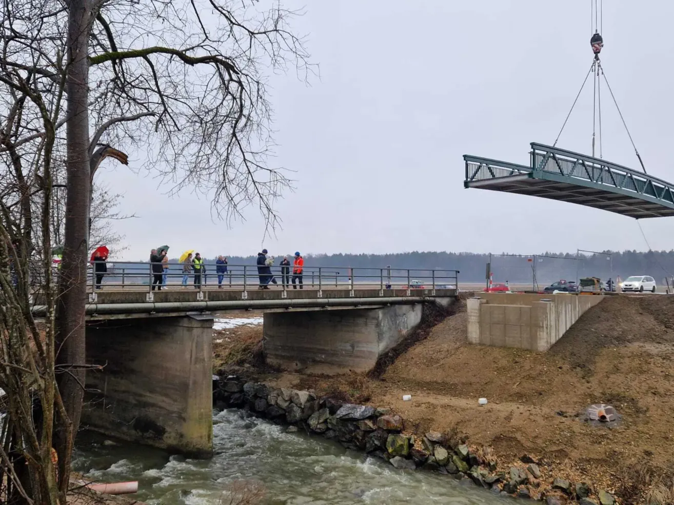 Eine Gruppe von Menschen steht auf einer Brücke über einem fließenden Fluss, einige halten Regenschirme. Rechts neben der Brücke ist ein Baukran zu sehen.