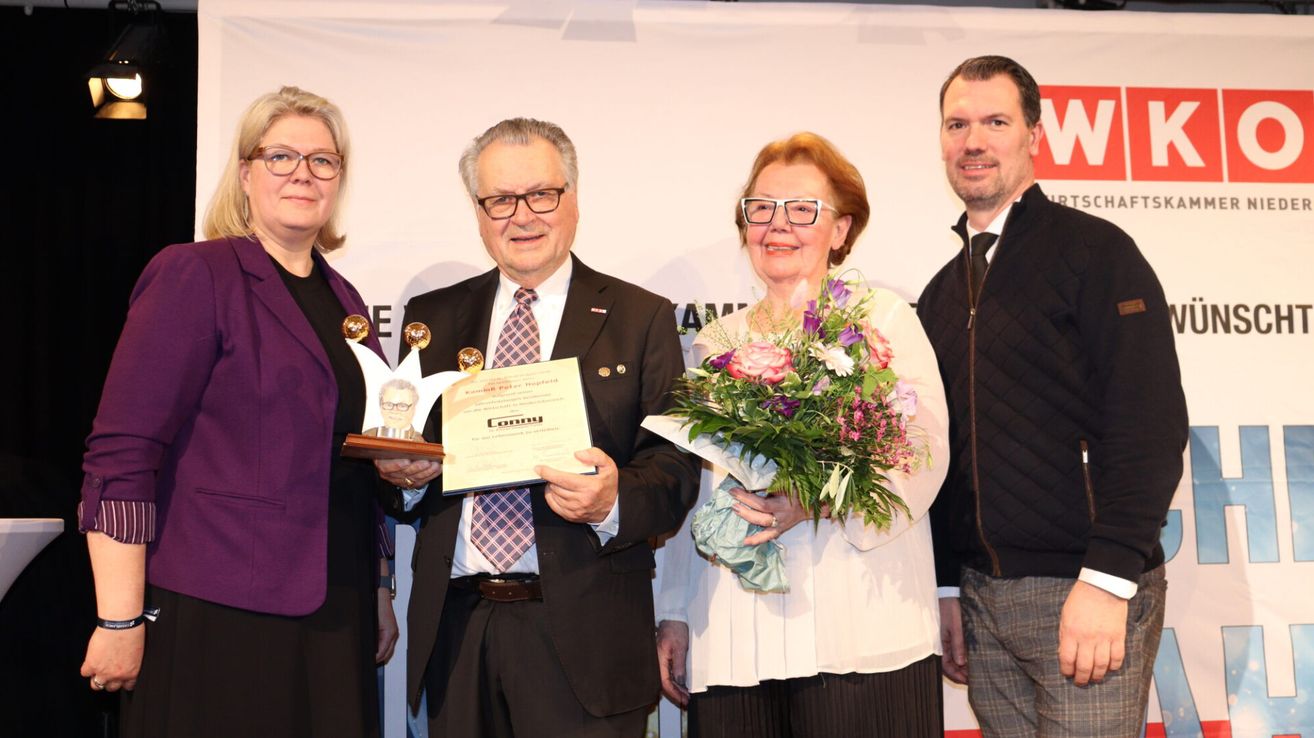 Vier Personen stehen für ein Foto, ein Mann mit Brille und Krawatte hält einen Preis. Neben ihm hält eine Frau einen Blumenstrauß. Dahinter steht eine weiße Kulisse mit Text.