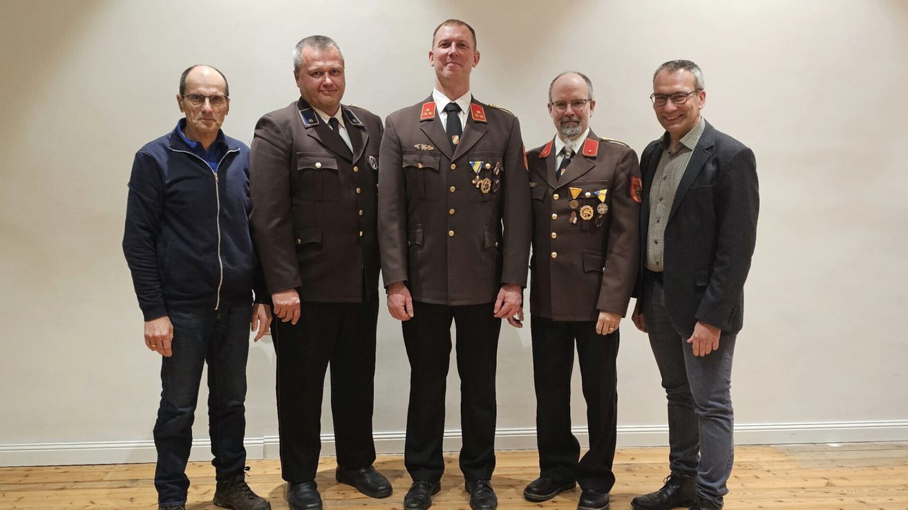 Fünf Männer stehen zusammen in einem Raum. Sie tragen alle Uniformen mit Medaillen und Krawatten. Der Mann in der Mitte lächelt und der Mann auf der rechten Seite schaut in die Kamera. Der Mann auf der linken Seite trägt eine blaue Jacke und schwarze Hose.