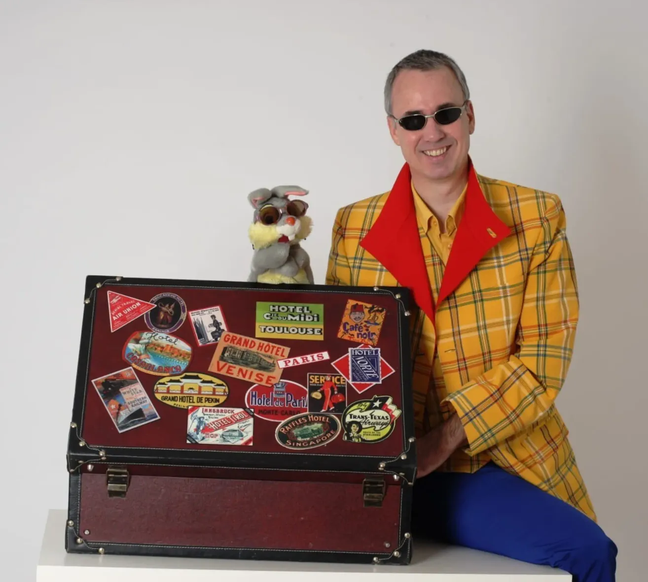 Ein Mann in einem gelben und grünen karierten Jackett hält einen mit Reiseaufklebern bedeckten Koffer. Er sitzt auf einer weißen Oberfläche mit einem Plüschtier hinter ihm.