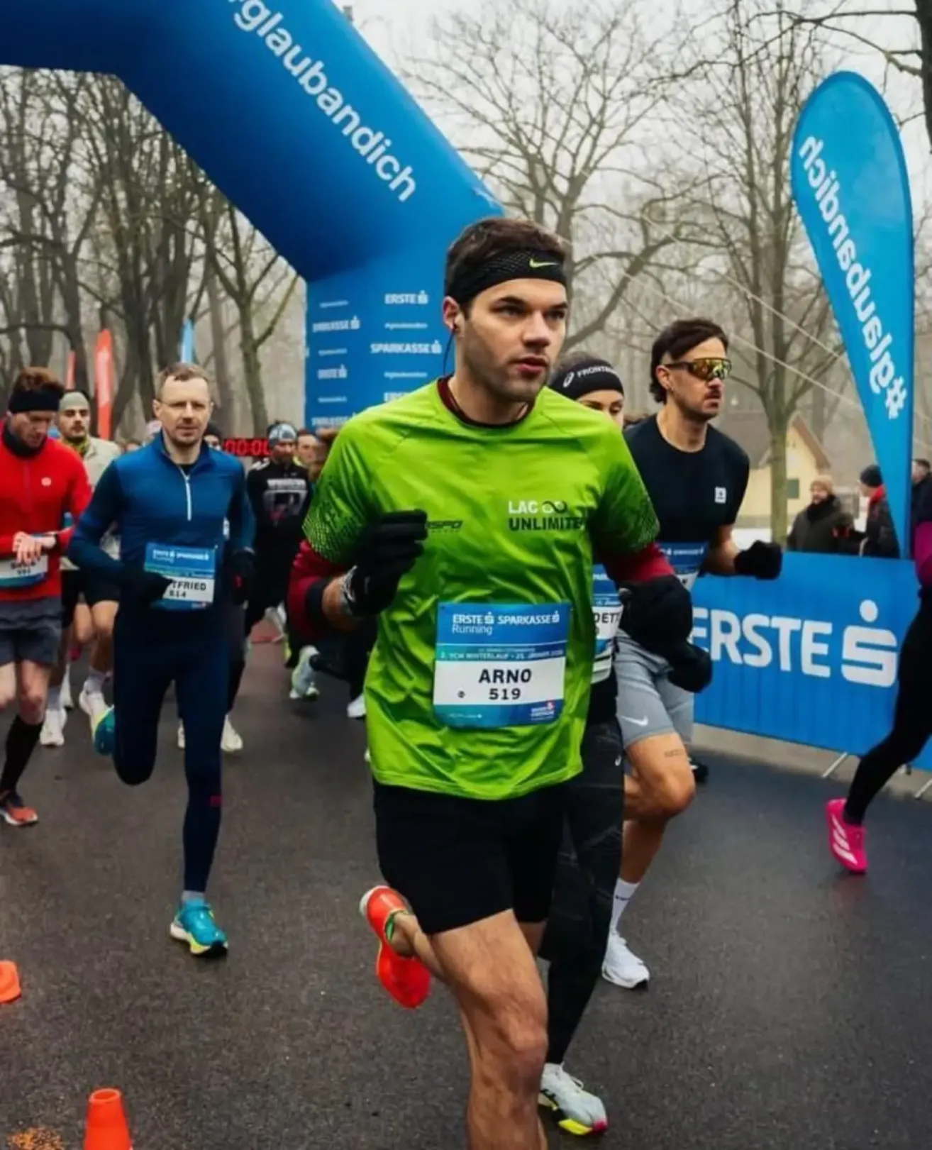 Läufer in verschiedenen Farben und Kleidung nehmen an einem Marathon teil. Der führende Läufer trägt ein grünes Shirt mit Lac Unlimite. Banner und Zuschauer sind hinter ihnen.