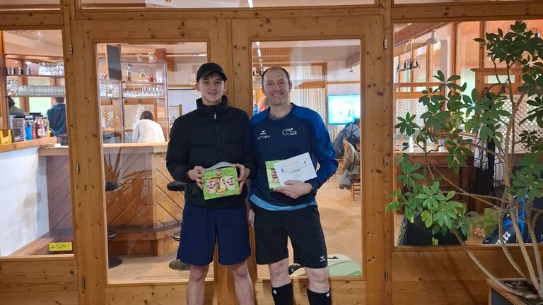 Zwei Männer stehen lächelnd vor einer Glastür. Einer hält eine Box und ein Zertifikat, der andere ein Zertifikat. Hinter ihnen steht eine Person in der Nähe einer Theke.