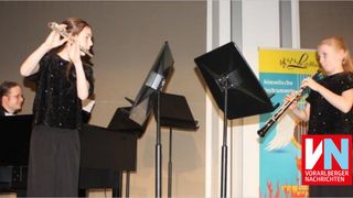 Zwei junge Musiker spielen auf der Bühne, einer spielt Flöte und der andere Klarinette. Hinter ihnen ist ein Klavier zu sehen, und vor ihnen stehen Notenständer. Auf der rechten Seite des Bildes ist ein Plakat mit Musiknoten zu sehen.