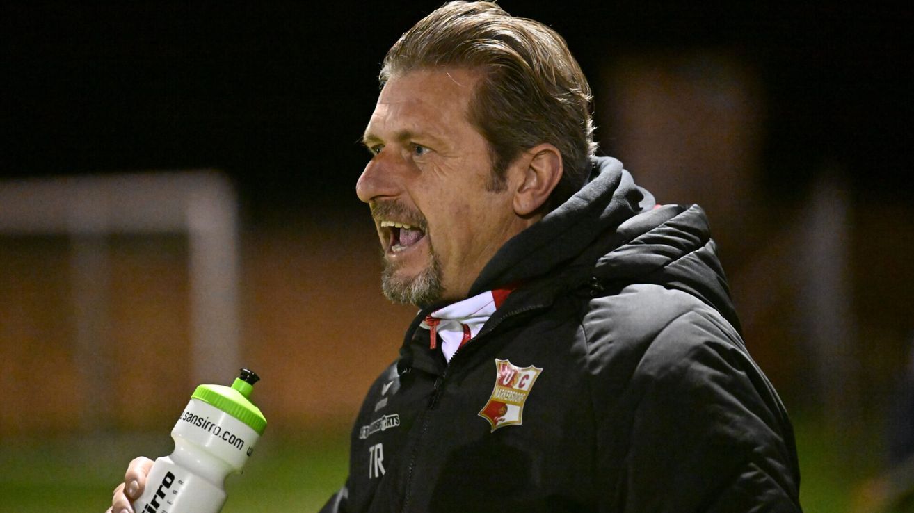 Ein Mann mit Bart und Schnurrbart lacht und trägt eine schwarze Jacke mit Logo. Links ist eine Flasche mit grünem Deckel und der Aufschrift siro.com zu sehen.