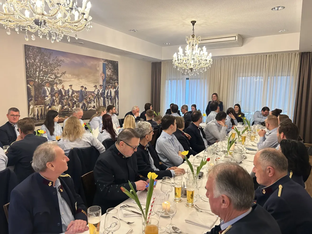 Eine Gruppe von Menschen in formeller Kleidung, die um einen langen Tisch herum sitzen, wahrscheinlich in einem Bankettsaal, mit Blumen, Kerzen und Weingläsern auf dem Tisch. Ein großer Bilderrahmen mit einem Bild von Soldaten ist an der Wand montiert.