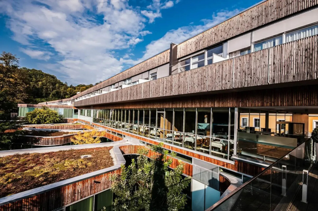 Ein modernes Gebäude mit Holzfassade und großen Glasfenstern, umgeben von Grün und einem blauen Himmel mit Wolken.