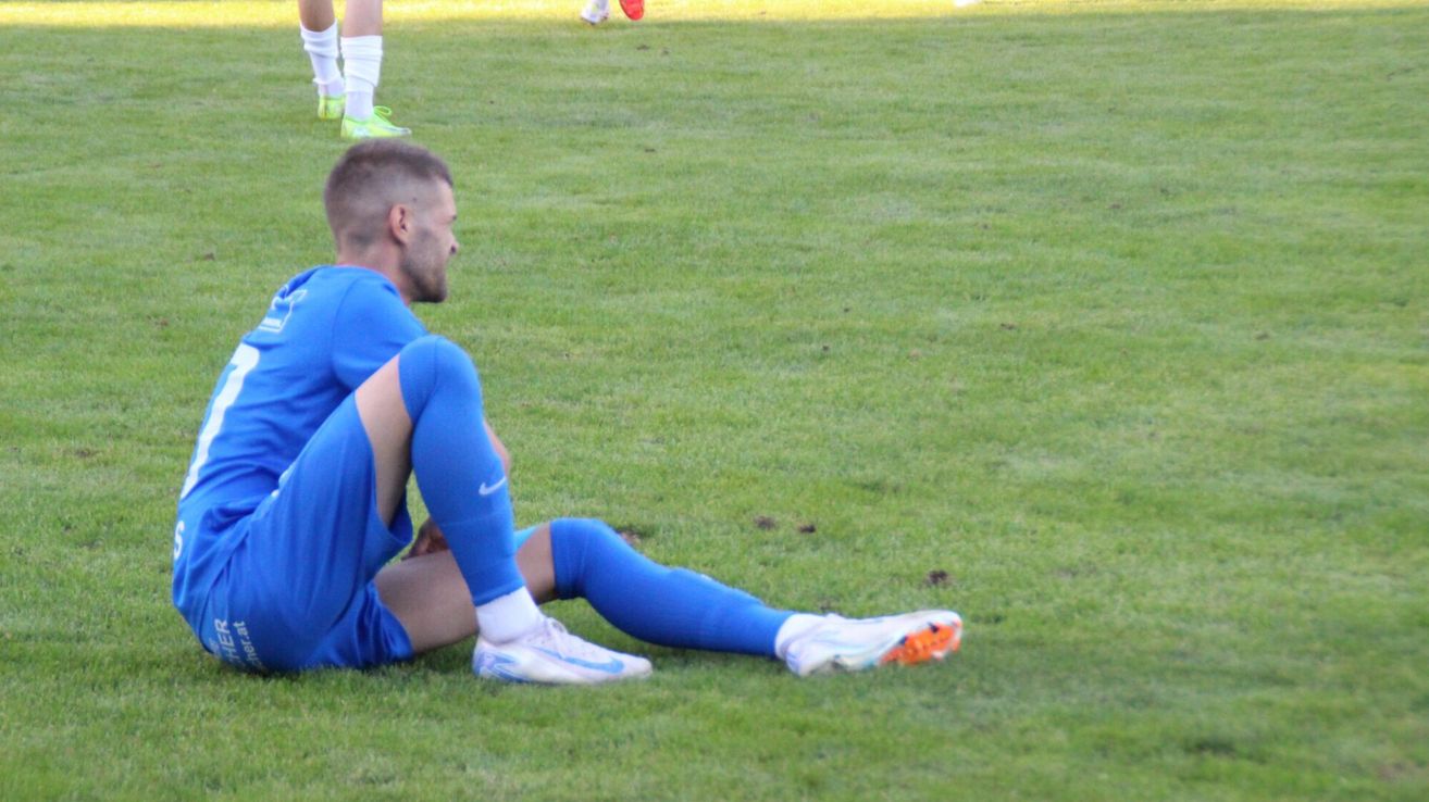 Ein Fußballspieler in einem blauen Trikot sitzt auf dem Rasen und schaut nach vorne, während andere in weißen Trikots hinter ihm stehen.