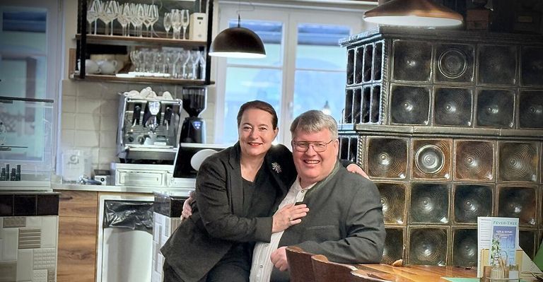Ein Paar posiert für ein Foto in einem Café, mit einem großen Fenster und Regalen mit Gläsern im Hintergrund.