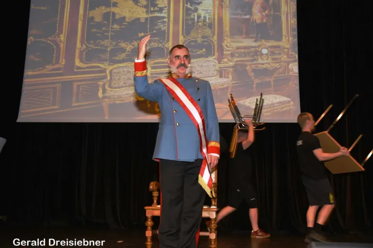 Ein Mann in einer blauen Uniform steht auf der Bühne und winkt mit der Hand. Eine Person steht hinter ihm mit einem Stuhl. Eine Projektionsleinwand befindet sich auf der Bühne.