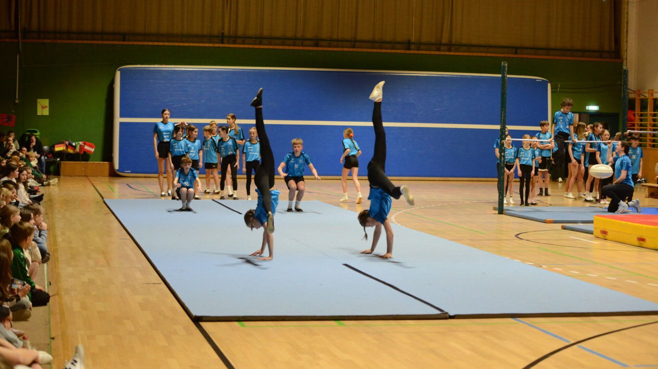 Eine Gruppe von Kindern in blauen Uniformen übt Gymnastik auf einer blauen Matte. Zwei Kinder machen Handstände, während andere zusehen. Der Ort ist drinnen mit Holzböden und einer blauen Wand.